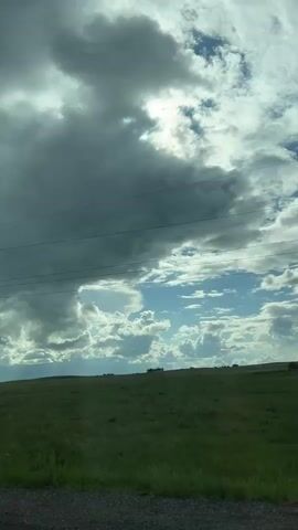A person records from inside a moving vehicle, capturing a panoramic view of a vast, green, undulating field stretching under a partially cloudy sky. A utility pole with power lines is visible on the left side of the frame initially. As the vehicle moves forward, the car's side mirror comes into view on the lower left, reflecting the arm and upper body of a person holding a smartphone to record the scene. The road shoulder is visible in the immediate foreground, next to the car.