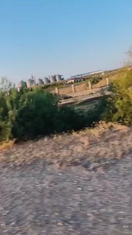The video shows a blurred view of an industrial area with large, cylindrical tanks. The tanks appear to be made of metal and are situated in an open, possibly rural, environment. There are also trees and bushes visible, along with some dry vegetation and gravel on the ground. The sky is clear and blue, suggesting daytime. The movement of the camera suggests it is mounted on a vehicle, possibly in motion.