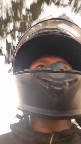 The video shows a close-up of a person wearing a black motorcycle helmet. The helmet is the primary focus, with the person's face partially visible through the visor. The background is blurred, indicating movement, and appears to be outdoors with trees and sky visible. The lighting suggests it is daytime. The camera angle is from the rider's perspective, looking forward.