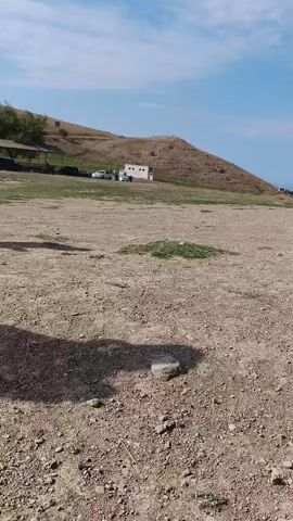 A dusty, barren, outdoor landscape with a gently sloping hill in the background under a bright blue sky with scattered clouds. A dirt and gravel ground covers the foreground and midground. A patch of green vegetation is visible in the midground. In the background, near the base of the hill, several cars are parked. A white building with a brown roof is also present. Some trees are on the left side of the hill. The camera is positioned low to the ground, possibly at ground level, and shows a shadow of an object or person in the foreground. The scene appears to be during daylight hours.