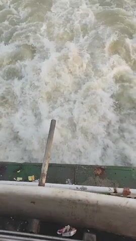 The video shows a churning body of water with white foam and turbid appearance, as seen from an elevated position above a concrete structure. Part of a rusted metal structure, possibly a fence or railing, is visible in the foreground, as well as what appears to be a wooden pole or post. A person's hand can be seen gripping the railing. Debris is scattered on the concrete platform. The water appears to be rapidly flowing, suggesting a dam or spillway.