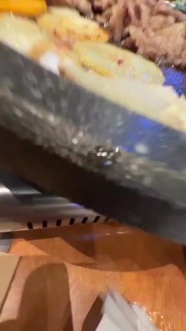 The video depicts a lively dining experience at a Korean BBQ restaurant. The scene opens with a close-up view of a large, round, dark-colored griddle placed on a portable gas stove. Various ingredients are cooking on the griddle, including sliced meats (resembling pork or beef belly), round slices of onions or potatoes, mushrooms, and lighter-colored items that appear to be seafood like shrimp or squid. Some yellow and blue pieces, possibly vegetables or other toppings, are also visible. A small white bowl containing a yellow liquid, likely egg yolk, sits beside the griddle.

The shot then widens to reveal a wooden table laden with numerous small metal dishes, commonly known as banchan, filled with various side dishes. A green bottle, possibly containing soju, and metal chopsticks are also on the table.

The video concludes with a focus on a young woman with long, light brown hair, wearing a white t-shirt. She displays a pronounced expression of surprise or excitement, with her mouth wide open and eyes wide. She holds a green leaf, likely a piece of lettuce for wrapping the grilled food. In the background, Korean characters on signs confirm the restaurant's cuisine, and a sign displaying "火鸡面" (huǒ jī miàn), meaning "fire chicken noodles," suggests the presence of spicy dish options. The indoor setting and lighting indicate that the meal is taking place during the evening.