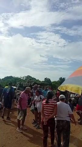 A bustling outdoor market scene unfolds under a partly cloudy sky. The ground is dirt and gravel, indicative of an unpaved area. In the foreground, a large, brightly colored umbrella, striped with orange, yellow, blue, and red, provides shade over a gathering of people. A man in a pink shirt and a woman in a white shirt are conversing near the umbrella. Further into the scene, more people are browsing stalls and interacting. Various vehicles, including trucks and motorcycles, are visible in the background, suggesting transport and commerce. Produce, such as corn and what appears to be root vegetables, are displayed for sale. People are seen walking, carrying bags, and engaging in transactions, creating a lively atmosphere of commerce and community. The overall impression is one of a typical open-air market on a bright, possibly warm day.