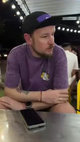 A man in a purple t-shirt and black hat sits at a metallic table. He is looking off to the side, then turns to face the camera. A black smartphone lies on the table. Other people are seated at tables in the background, under string lights. Yellow plastic chairs surround the tables.