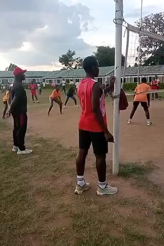 The video shows a group of people playing volleyball on a dirt court. The players are wearing different colored athletic tops and shorts. There is a volleyball net in the center of the court. One person in a red tank top stands near a net pole, watching the game. There are trees and a building in the background. The sky is overcast.