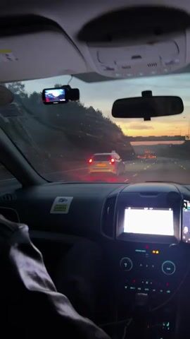 The video shows the interior of a vehicle driving on a road at dusk or dawn. The view is from the perspective of a passenger looking forward through the windshield. The dashboard is visible, with a navigation screen on the right and a media display on the left showing song information. A dash camera is mounted on the windshield. Outside, there is a highway with other vehicles, and the sky displays a gradient of orange and blue. Trees line the sides of the road, and streetlights are visible in the distance. The lighting suggests it is either early morning or late evening.