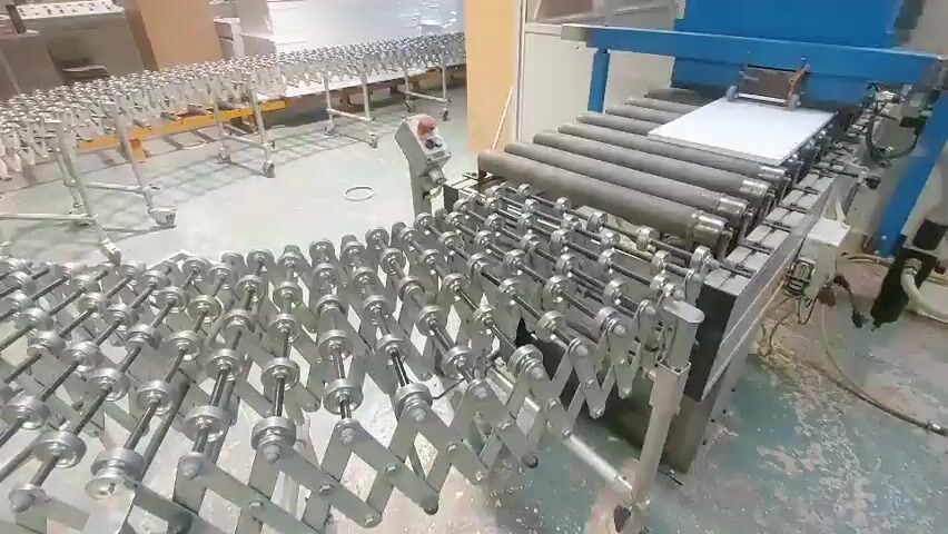 This video captures an industrial setting where a white rectangular panel is being transported along a conveyor belt system. The system consists of multiple roller conveyors, some of which are articulated and extendable, creating a flexible pathway. The panel is moving from right to left in the foreground and then curves around a bend. In the background, a more extensive roller conveyor system is visible, with many more panels or materials stacked on it. The environment appears to be a factory floor with concrete flooring and industrial machinery. There are no people visible in the video. A red and white warning sign is attached to one of the articulated conveyors.