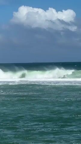 The video shows a wide shot of the ocean on a bright, partly cloudy day in Walgama, Sri Lanka. Large green and white waves continuously roll and break across the frame. The water is a deep teal color closer to the foreground, becoming a darker blue further out. The sky above is a clear blue with several large, white and grey clouds scattered across it. No people or artificial objects are visible. The scene captures the natural movement and power of the ocean waves under a sunny sky.