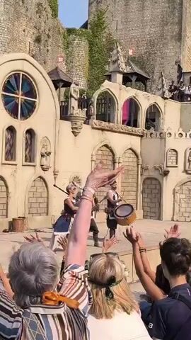 The video shows a performance taking place in front of a medieval-style castle backdrop. In the foreground, a crowd of spectators is visible from behind, with their arms raised in the air. On a raised platform in the middle ground, a group of performers are on stage. One performer, dressed in a blue and white outfit, plays bagpipes. Another performer, wearing a brown tunic and breeches, strums a lute. A third performer, in similar medieval attire, beats a large drum. The stage itself is decorated with stone-like textures and arched windows. Behind the stage, the castle structure rises, featuring crenellated walls, banners, and turrets. Sunlight suggests the time of day is daytime. The overall atmosphere appears to be that of a historical reenactment or festival.