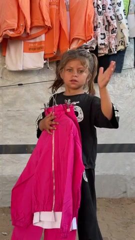 A young girl with pigtails, wearing a black shirt and pants, stands in front of racks of clothing. She holds a pink jacket and pants on hangers. She is waving at the camera. The clothing racks contain various articles of clothing, including orange jackets and patterned shirts. The background appears to be outdoors, possibly a market stall.
