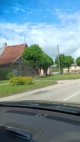 The video shows a front-facing view from inside a car driving through a town square. The sky is partly cloudy and blue. On the left side of the road, there are several green trees, and beyond them, old buildings with red and brown roofs are visible. Some cars are parked along the street and in the distance. The road ahead curves to the right, and a few street signs or kiosks are on the right side of the road. The car's dashboard and windshield are visible in the foreground.