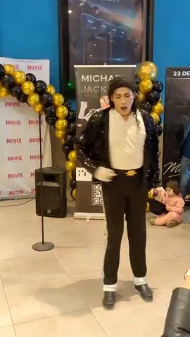 A male performer dressed as Michael Jackson, wearing a black sequined jacket, white shirt, black pants, white socks, and black shoes, is dancing in front of a banner that says "MICHAEL JACKSON". Gold and black balloons are arranged in an arch to the left of the performer. A speaker on a stand is to the left of the balloons. In the background, there is a movie poster. To the right of the banner, there are more gold balloons and a section of a wall with text that appears to be a date. Behind the performer and to the right, a man and a child are visible. The floor is tiled. The lighting appears to be artificial. The performer is in motion, with his mouth open as if singing or shouting.