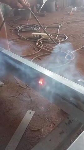 A person is welding a metal beam. The person is holding a welding torch in their right hand and a metal rod in their left hand. The torch is emitting sparks and smoke, and the metal beam is glowing red where it is being welded. There are several electrical cords and a power strip on the ground in the background. The ground is made of dirt.