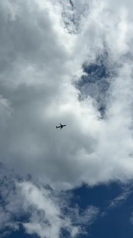 A passenger jet airplane is visible in the sky, silhouetted against a backdrop of clouds and a deep blue sky. The airplane is positioned in the center of the frame, flying from left to right. Large, puffy white clouds dominate the upper and left portions of the image, with patches of darker blue sky visible through breaks in the clouds. The airplane appears to be at a considerable altitude. The overall impression is one of a clear, bright day with scattered cloud cover, and the airplane is in flight.