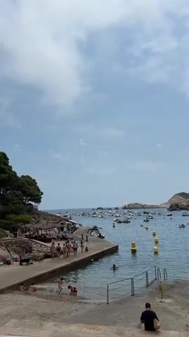 The video shows a busy beach scene with people enjoying the water and the coastline. In the foreground, a concrete walkway slopes down to the water, with several children playing near the edge. Further along the walkway, a group of people are standing and talking. A concrete pier extends into the water, with more people gathered on it, some appearing to be sunbathing or relaxing. The water is calm and clear, with several yellow buoys marking a swimming area. Numerous boats are anchored in the bay, and in the background, rocky islands rise from the sea. Lush green trees and vegetation cover the hillside to the left of the scene. The sky is partly cloudy. One person is seated on the lower foreground steps, facing away from the camera.