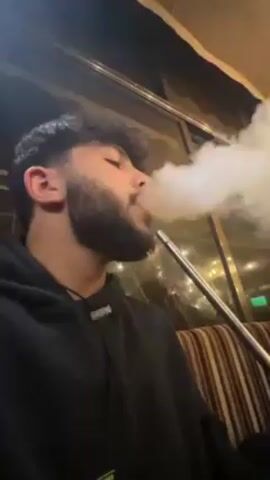 A young man with a beard is sitting in what appears to be a lounge, smoking a hookah. He is wearing a black hoodie. The background features a window with lights visible outside. The man takes a drag from the hookah, exhaling a large cloud of smoke. The music plays in the background. The camera slightly pans around the man.