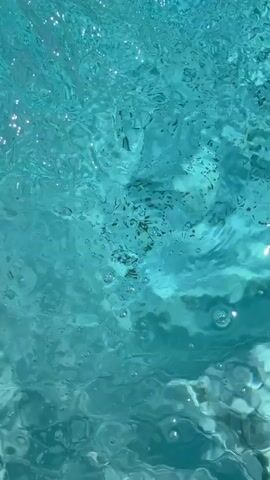 The video shows a close-up view of turquoise water from below the surface. Bubbles are rising, and light glints off the water's surface, creating a shimmering effect. The water is disturbed, indicating movement, possibly from a person or an object within the pool. The bottom of the pool is visible in some areas, showing white rectangular tiles.