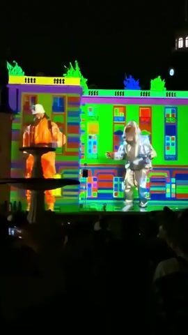 The video shows a projection mapping show on a building at night. The projections feature abstract colorful patterns and animated figures of people. The figures appear to be dressed in brightly colored work-related outfits, like construction workers or firefighters. They are shown in various poses, some dancing or interacting with the projected environment. In the foreground, silhouetted audience members are visible watching the show. The overall effect is a dynamic and colorful visual performance.
