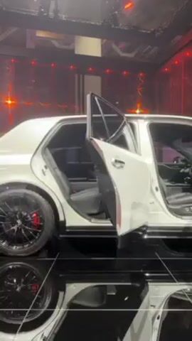 The video shows a white sedan with black rims displayed in what appears to be an exhibition hall. The car doors on one side are open, revealing the interior. The car is elevated on a black reflective platform. In the background are red lights and a patterned design. No people are visible.