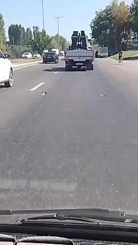 The video shows a view from inside a car driving on a paved road with multiple lanes. The road is lined with trees and streetlights. Several vehicles are visible on the road, including a white SUV in the left lane, a black car in the middle lane, and a flatbed truck carrying a large black object in the right lane. The truck appears to have its hazard lights on, and its rear doors are open. The weather is clear and sunny.