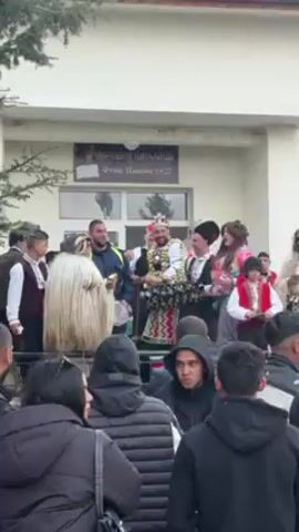 A crowd of people has gathered in front of a two-story building with a sign above the door. The sign reads "HAPOAH ЧИTAAHHЩE Oтец Паисий 1927". A group of people are standing and dancing on the building's porch, including at least one child. The dancers are dressed in traditional clothing and furry costumes. A uniformed police officer is standing on the porch near the dancers. One of the dancers, a woman wearing a red dress, walks up to the officer and puts her arms around him as though giving him a hug.