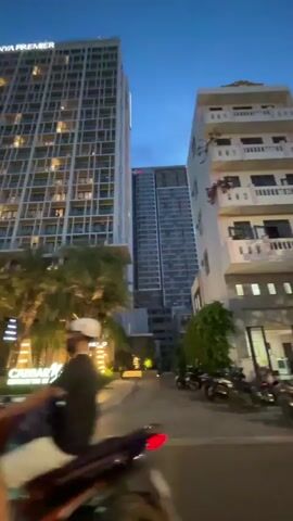 The video shows a wide shot of an urban street scene at dusk. In the foreground, there is a road with a few parked motorcycles on the right. Across the street, a well-lit entrance area is visible, featuring palm trees and decorative lighting. A prominent sign reads "CABBAR NOTTE" in illuminated letters. Behind this, a tall, modern apartment building with many lit windows stands out, displaying the text "ANYA PREMIER" near its top. To the right of the "Anya Premier" building, another, taller skyscraper is partially visible, and further to the right, a white building with multiple balconies can be seen. The sky is a deep blue, indicating twilight.