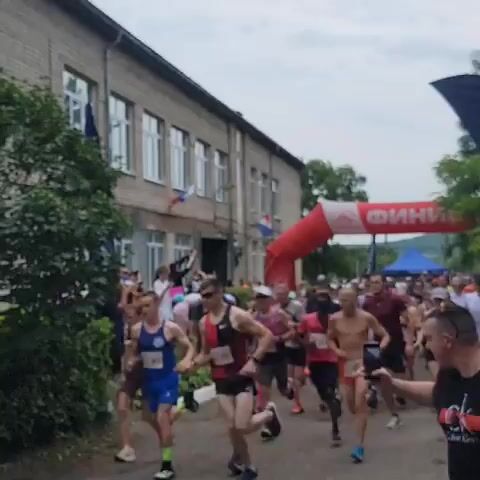 The video shows a group of runners in a race, moving from left to right across the frame. They are running on a dirt path, with a building and trees in the background. There is a large red inflatable archway with the word "ФИНИШ" (FINISH) visible on it, indicating the end of the race. The runners are wearing various athletic clothing, including shorts, t-shirts, and running shoes. Some runners have bib numbers attached to their shirts. The weather appears to be overcast.