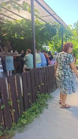 The video opens with a wide shot of an outdoor seating area under a canopy of green vines. Several people are seated at tables, some engaged in conversation. A woman with red hair wearing a floral dress stands to the right of the frame, facing away from the camera. She is holding a white bag. Behind her, another woman with blonde hair, wearing a light blue shirt and denim shorts, is also facing away from the camera. The scene is outdoors with trees and foliage visible in the background. The ground is a paved walkway. The video appears to be a short, informal recording of a gathering.