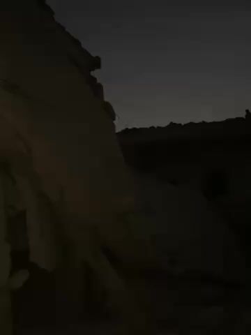 The video shows the ruins of buildings in dim lighting, possibly at dusk or dawn. The buildings appear heavily damaged, with sections collapsed or missing, revealing their interiors. Debris and rubble cover the ground. The atmosphere is somber and conveys a sense of destruction. There are no people visible in the video.