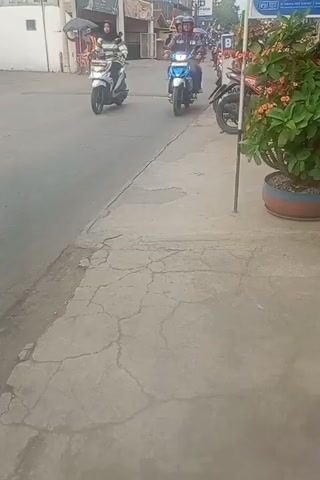 The video shows a street scene from a first-person perspective, likely from a moving vehicle or person. The focus is on a road with several motorcycles passing by. On the left side of the road, a white scooter is being ridden by a person wearing a hijab and a striped jacket. Further down the road, a blue and white motorcycle with two occupants, both wearing helmets, is also visible. To the right of the road, there is a sidewalk with a potted plant and a pole. More motorcycles are seen in the background, suggesting a busy street. The ground is paved with cracked concrete. The overall impression is a typical urban street scene with motorcycle traffic.