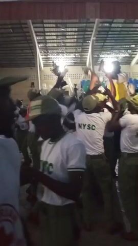 A group of young people in white t-shirts with "NYSC" printed on the back and green trousers are gathered in a semi-covered outdoor area. They are raising their arms and cheering, some holding up what appears to be a trophy or award. The lighting suggests it is either dusk or early morning. The environment seems to be a communal gathering space, possibly a camp or event venue. The overall mood is celebratory and energetic.