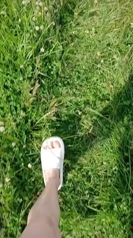 A first-person perspective video shows a person walking through a grassy area. The person is wearing white slides and is walking on a path covered in green grass and small white flowers, possibly clover. The camera angle is looking down at the person's feet and the ground. The scene appears to be outdoors on a sunny day, with shadows indicating sunlight.