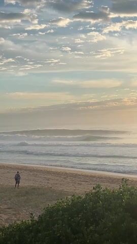 The video shows a beach scene at dawn or dusk. A person walks on the sand near the vegetation. In the background, waves roll in towards the shore. The sky is filled with clouds, and the sun casts a soft glow over the scene.