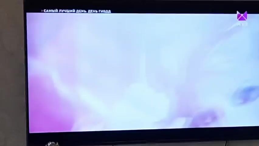 The video captures a television screen displaying a music video. The scene is dimly lit with a predominant blue hue. The top left corner of the screen shows Russian text, possibly a song title or artist name, "САМЫЙ ЛУЧШИЙ ДЕНЬ ГИБДД". In the top right corner, a purple logo with the letters "МУЗ" is visible, indicating the channel.

The video begins with a close-up of a person's face, focusing on their mouth and neck, with a blue light casting a glow. As the video progresses, the scene shifts to reveal a group of people dressed in dark clothing, possibly uniforms, with one person in the foreground wearing what appears to be a police-style hat and jacket. The group is in motion, with some individuals gesturing and dancing. The overall aesthetic suggests a dramatic and possibly theatrical performance. The camera work is dynamic, with quick cuts and close-ups.