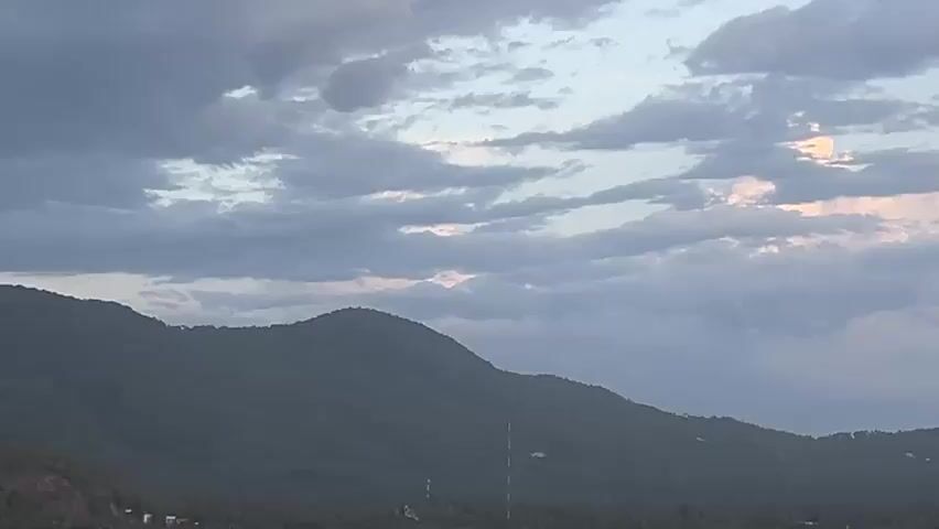 The camera pans out from a mountain range to reveal a view of the ocean with an island in the distance. The sky is cloudy with streaks of pink and orange light. In the foreground, a tree branch with leaves is partially visible in the upper right corner of the frame. The overall scene depicts a landscape view at what appears to be dusk or dawn.