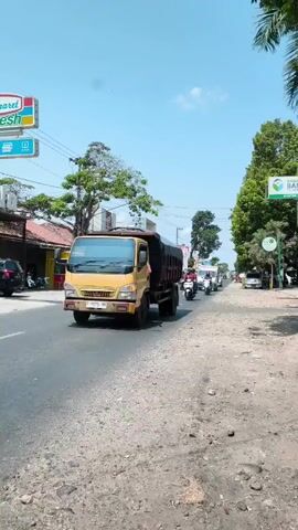 A bright, sunny day with a clear blue sky. A yellow dump truck drives down a paved road. To the left, there are shops and signs, including one for "Indomaret Fresh." To the right, there is a dirt shoulder with rocks and sparse vegetation, leading to trees. Several motorcycles and a white van are also on the road, moving in the same direction as the dump truck. The scene appears to be a typical street in a town or village.