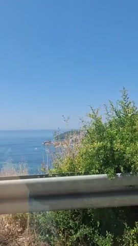 The video shows a view of a coastline from a moving vehicle. In the foreground, there is a metal guardrail and some dry vegetation. Beyond the guardrail, a body of blue water stretches to the horizon, dotted with several white boats. Along the coast, a town with red-tiled roofs and white buildings is visible, nestled at the base of a green, hilly landscape. The sky is clear and blue. The camera appears to be panning slightly as the vehicle moves.