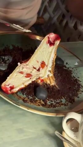 The video shows a slice of strawberry cheesecake on a plate with chocolate crumble and dark gray garnishes. A fork is actively being used to eat the cheesecake. There is a knife on the plate as well, and a white cup is visible in the lower right corner. The background appears to be a restaurant setting with a blurred person wearing a white shirt.