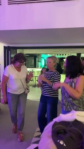 The video features three middle-aged women dancing indoors. The woman on the left wears a white t-shirt, grey pants, and has short brown hair. The woman in the middle wears a striped black and white shirt and jeans, and has blonde hair pulled back in a bun. The woman on the right wears a printed sleeveless top and jeans. They are dancing to music, and appear to be enjoying themselves. The room is lit with purple light, and a doorway leads to an outdoor area with green lighting. Behind the women are some seated people. In the foreground is a pile of clothing.