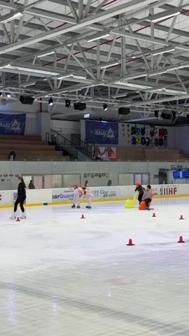 The video shows an indoor ice skating rink. Several individuals are present on the ice, including children and adults. The subjects are engaged in various activities: some are figure skating, one person appears to be teaching or supervising, and children are using assistive devices for skating, such as a blue dolphin-shaped sled and a yellow walker.

There are several orange traffic cones placed on the ice, likely for drills or to mark a course. In the background, there are bleachers with some spectators seated. Banners with "ISRAEL" written on them are visible hanging from the ceiling. Logos for "FIGURE SKATING ACADEMY ISRAEL" and "IIHF" are also visible on the rink's boards.

The lighting suggests it is daytime, but the interior setting makes the time of day less definitive. The overall atmosphere appears to be one of learning and recreation. There is no indication of weather conditions as it is an indoor facility. The location is confirmed as Qalansawe, Israel, by the provided context.