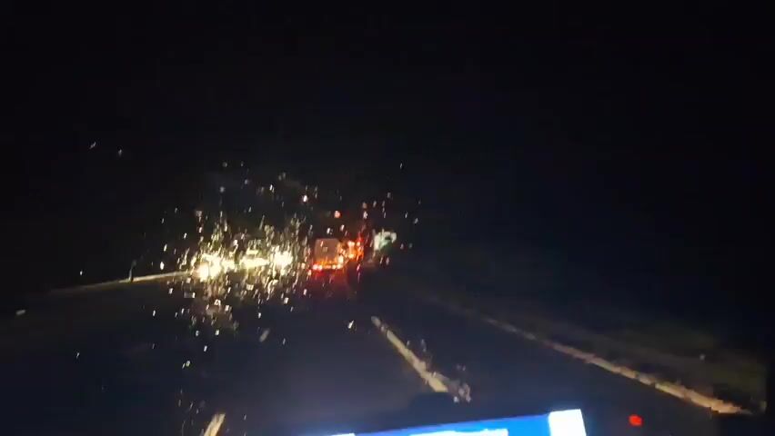 The video shows a nighttime view from inside a vehicle on a road. Sparks are visible on the left side of the frame, appearing to originate from the road or the side of the road. In the distance, there are several vehicles with their brake lights illuminated, indicating traffic congestion or a stopped vehicle. One vehicle, possibly a truck, is clearly visible with red brake lights. A highway sign is also visible in the distance on the right. The road has white lane markings. The overall impression is of a nighttime drive on a highway with an unusual spectacle of sparks and slow-moving traffic ahead.