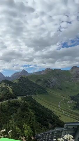 The video pans across a lush green mountain landscape under a partly cloudy sky. The foreground shows the top edge of some solar panels and greenery, possibly flowers or small plants. The middle ground features rolling green hills with winding roads, scattered trees, and a small village with red-roofed buildings nestled in a valley. In the background, jagged mountain peaks rise against the sky. The camera moves slowly, revealing more of the vast mountainous terrain. There are no people visible in the video.