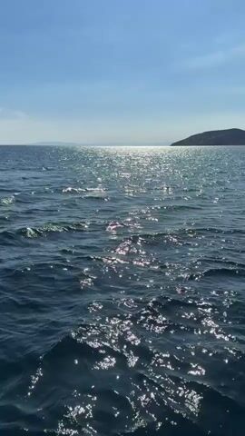 The video captures a wide shot of a calm, deep blue sea under a clear, bright blue sky. The sun is high, creating intense sparkling reflections on the water's surface, forming a shimmering path towards the horizon. Gentle waves and ripples are visible, reflecting the sunlight in dynamic patterns. In the distance, on the right side of the frame, a dark landmass, likely an island or coastline, can be seen. The horizon line is distinct, separating the vast expanse of water from the cloudless sky. The scene evokes a peaceful, sunny day on the water, consistent with the coastal location of Palea Fokea Municipal Unit, Greece. No people or specific objects are prominently featured, and there is no visible text.