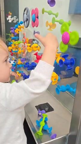A young child with blonde hair is playing with colorful suction cup toys on a reflective surface. The child, wearing a light grey long-sleeved shirt, stands in front of the surface, which is covered with various suction cup toys in an array of colors. The child is actively sticking and arranging the toys on the reflective surface, which appears to be a part of an exhibit or play area. Some suction cup toys are also on the countertop in front of the mirror.