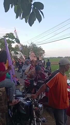The video shows a group of people gathered around motorcycles on a road in a rural area. Some individuals are seated on the motorcycles, while others stand nearby. In the background, there's a decorated structure, possibly part of a parade or procession.  A young girl sits on a concrete barrier to the left, holding purple foam tubes.  The people in the scene appear to be dressed casually. The environment includes trees, a rice field, and overhead power lines. The sky is visible, suggesting the scene is taking place outdoors during daylight.