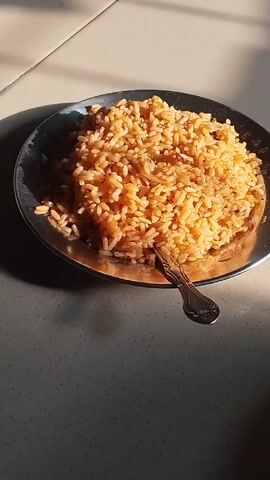 A plate of rice sits on a table, illuminated by sunlight. A spoon rests on the side of the plate. Music plays in the background.