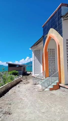 The video shows a bright blue sky over a body of water and mountains. In the foreground, there is a white building with an orange archway and stairs leading up to it. The building appears to be under construction or recently completed. Beyond the building, there are several small structures and vegetation, with the water and mountains in the distance. The water is calm and reflects the sky. There are clouds in the sky and on the mountains. The overall scene is peaceful and scenic.