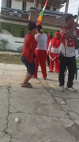 The video shows a group of people in an outdoor setting, possibly a courtyard or paved area in front of a building. In the foreground, a person wearing a red shirt and blue shorts is facing away from the camera. To their right, another person wearing a white shirt and red pants is also facing away. Behind them, several individuals are visible, including two women wearing red hijabs and matching red pants. One of the women is laughing and gesturing with her hands. Another person, dressed in a white and red jersey with the number 80, stands to the far right, holding what appears to be a microphone or phone. A flag with red, yellow, and green stripes is visible attached to a pole in the background, near the building. The ground is paved with cracked concrete. The overall atmosphere seems to be one of lightheartedness or a casual gathering.