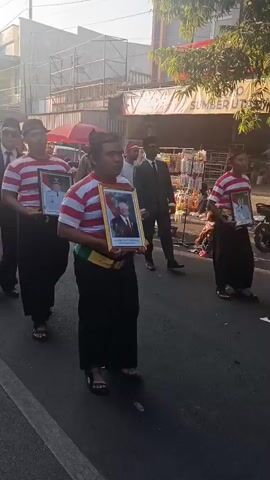A group of people are marching in a procession on a paved road. In the foreground, three individuals are walking from left to right. The first person on the left is wearing a dark suit, white shirt, and red tie, with sunglasses and a cap. To their right, a person in a red and white striped shirt and black pants holds a framed portrait of a man in a uniform. Another person in a similar red and white striped shirt and black pants, wearing a green sash and a red head covering, holds a framed portrait of a man in a suit. Further back, more people are visible, some in traditional clothing and others in formal attire. The procession appears to be moving down a street lined with buildings and shops, with some onlookers on the right side of the road. The sky is bright, suggesting it is daytime.