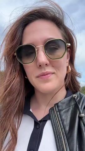 A woman in sunglasses and a black leather jacket is hiking on a gravel path in a mountainous area. She is facing the camera and holding it, creating a selfie video. Other hikers are visible in the background, walking in the same direction. The landscape includes grassy slopes and scattered trees. The sky is partly cloudy. There is a slight breeze, causing her hair to blow.